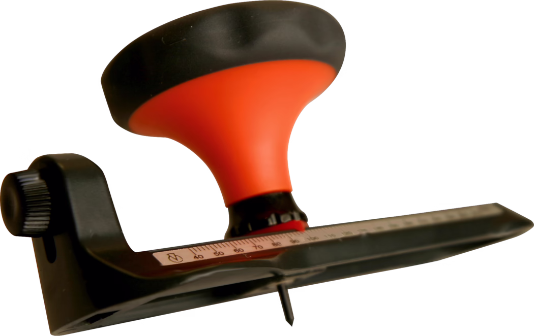 WOLFF_Dummy_15624_circular cutter_2025-08 Close-up of a handheld woodworking tool (possibly a sander or plane) featuring a bright orange ergonomic handle with a black top. A black plastic adjustment knob is visible on the side and a metal ruler with engraved numbers and markings runs along the base. A pointed screw or blade protrudes from the bottom. The tool is shown isolated on a white background with shallow depth of field that emphasizes the handle and measuring scale while other parts are softly out of focus.