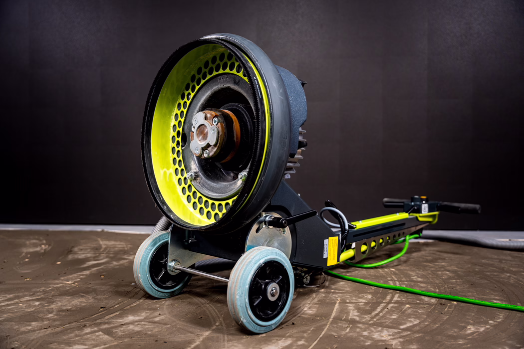 WOLFF_Application Image_Mambo_2024-01 (18) A heavy-duty floor sanding/grinding machine photographed in a studio setting. The image shows a large circular sanding head with a perforated bright yellow-green inner ring and a central metal hub with exposed bolts and some surface rust. The machine has a sturdy dark gray metal frame, two light-blue caster wheels in front, a long handle with black grips and control levers at the rear, and a green power/hose cable coiled on the floor. The concrete floor around the device is dusty with visible sanding marks. The lighting highlights the machine's textures—metal, rubber and worn paint—against a plain dark backdrop.