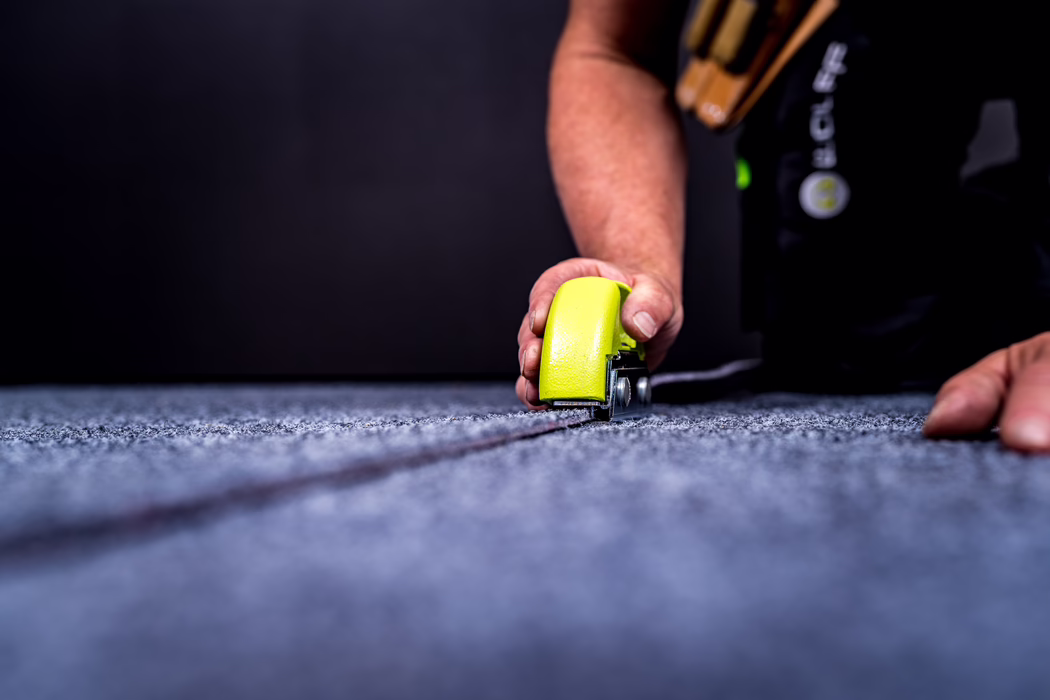 WOLFF_Application Image_GreenCut_Detail2_2022-11 Close-up of a worker's hand using a yellow carpet tool to trim and join a grey carpet seam; shallow depth of field focuses on the tool and seam while the background is dark and out of focus. The scene highlights manual flooring work, precision and the texture of the carpet fibers.