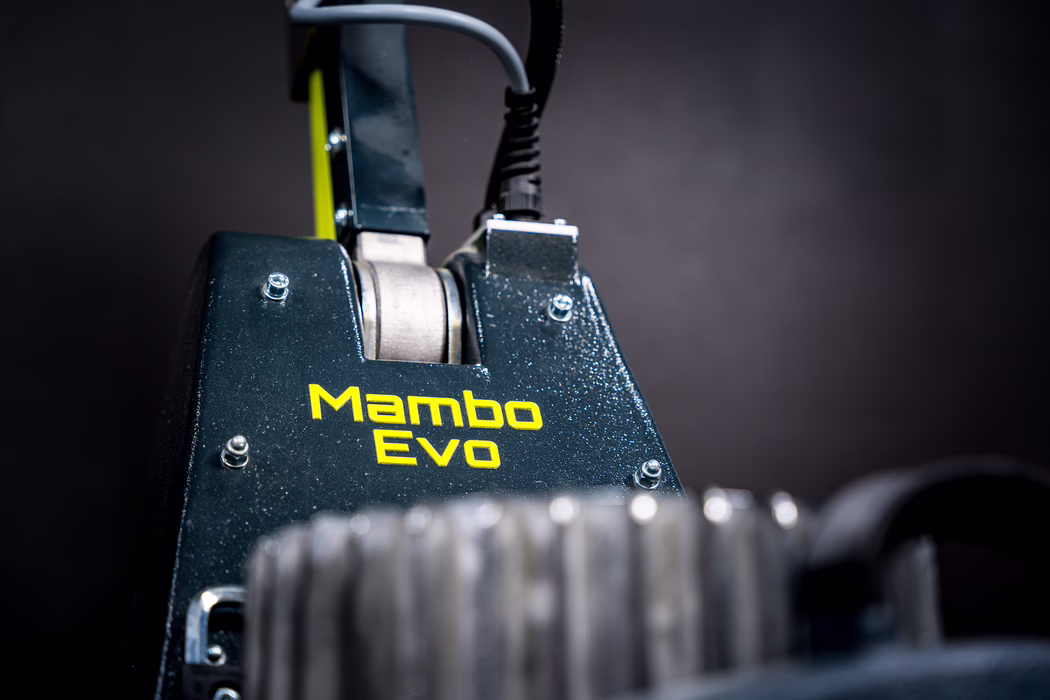 WOLFF_Application Image_Mambo_2024-01 (11) Close-up of a dark metallic machine component with the bright yellow "Mambo Evo" logo on a textured surface. Screws, a coiled cable and a metal roller are visible; the foreground is blurred and the background is a dark studio backdrop, giving a shallow depth of field and an industrial, technical feel.